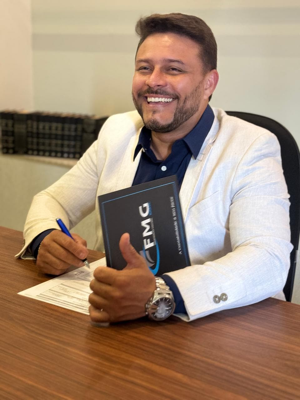 Adriano Silva, sócio-fundador da FMG Brasil, sorrindo à mesa do escritório com uma pasta da FMG
