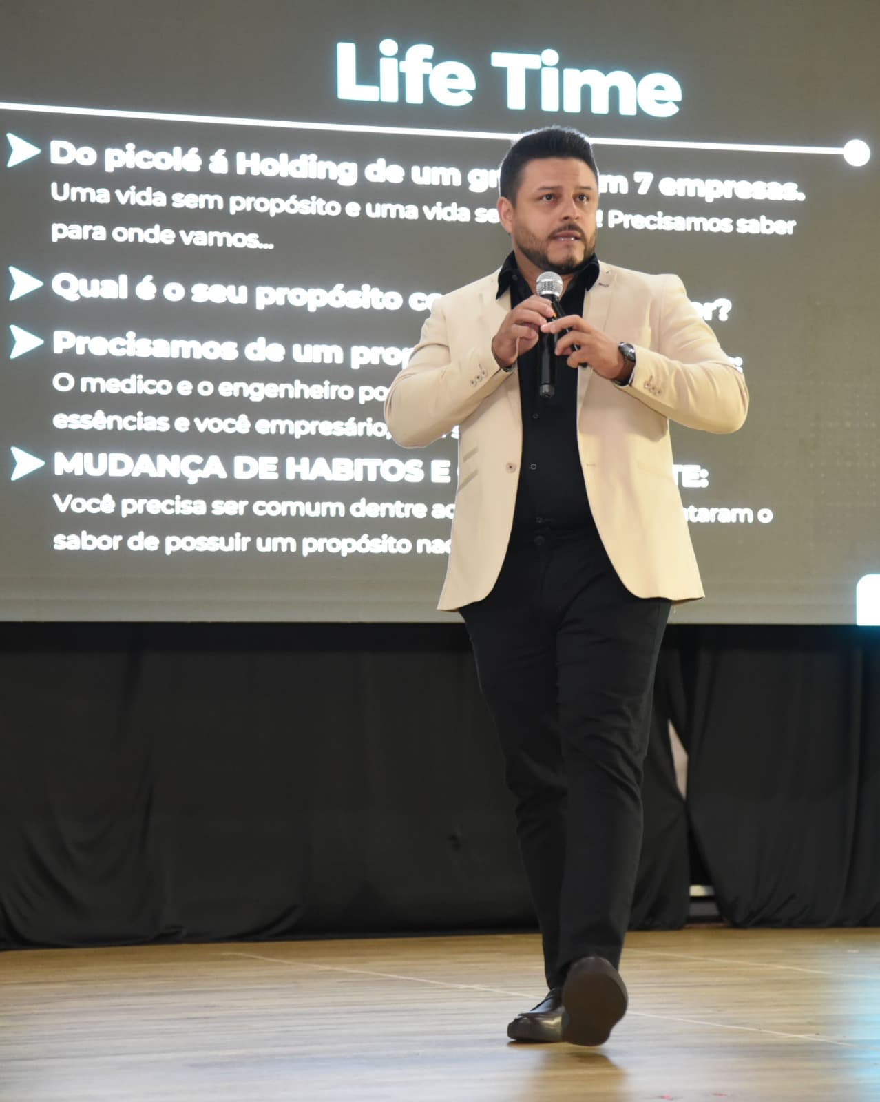 Adriano Silva palestrando no palco, com microfone, diante de um telão